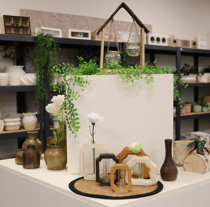 A display featuring small wooden vases with white flowers and trailing greenery, geometric wooden objects with small glass tubes on a natural fiber placemat, stacked books, and a decorative hanging terrarium with plants on a white surface, set against dark open shelving with ceramics